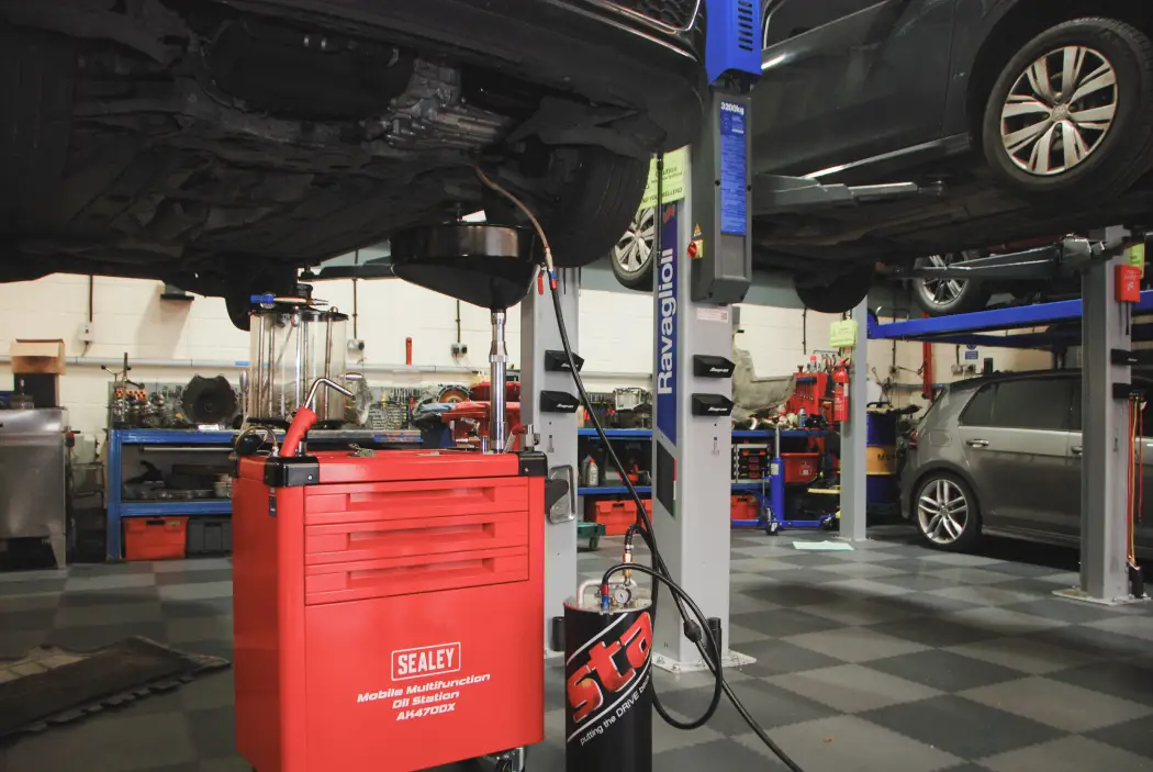 A DSG gearbox service taking place in a professional workshop, showing a car lifted on a hydraulic ramp with oil draining equipment and a red Sealey mobile oil station underneath. The workshop is clean and well-equipped with tools, lifts, and vehicles in the background.